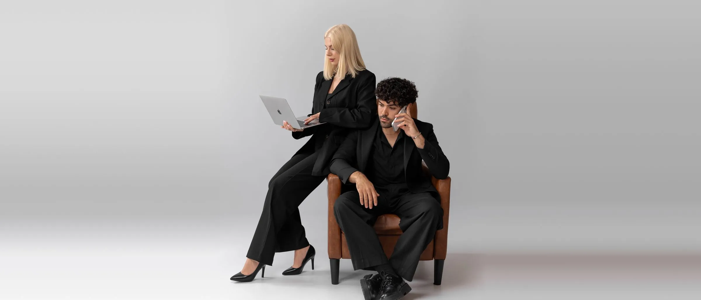 Nikola Miller working on a laptop beside Gui Ferreira on a phone call, seated in a leather armchair in a grey studio