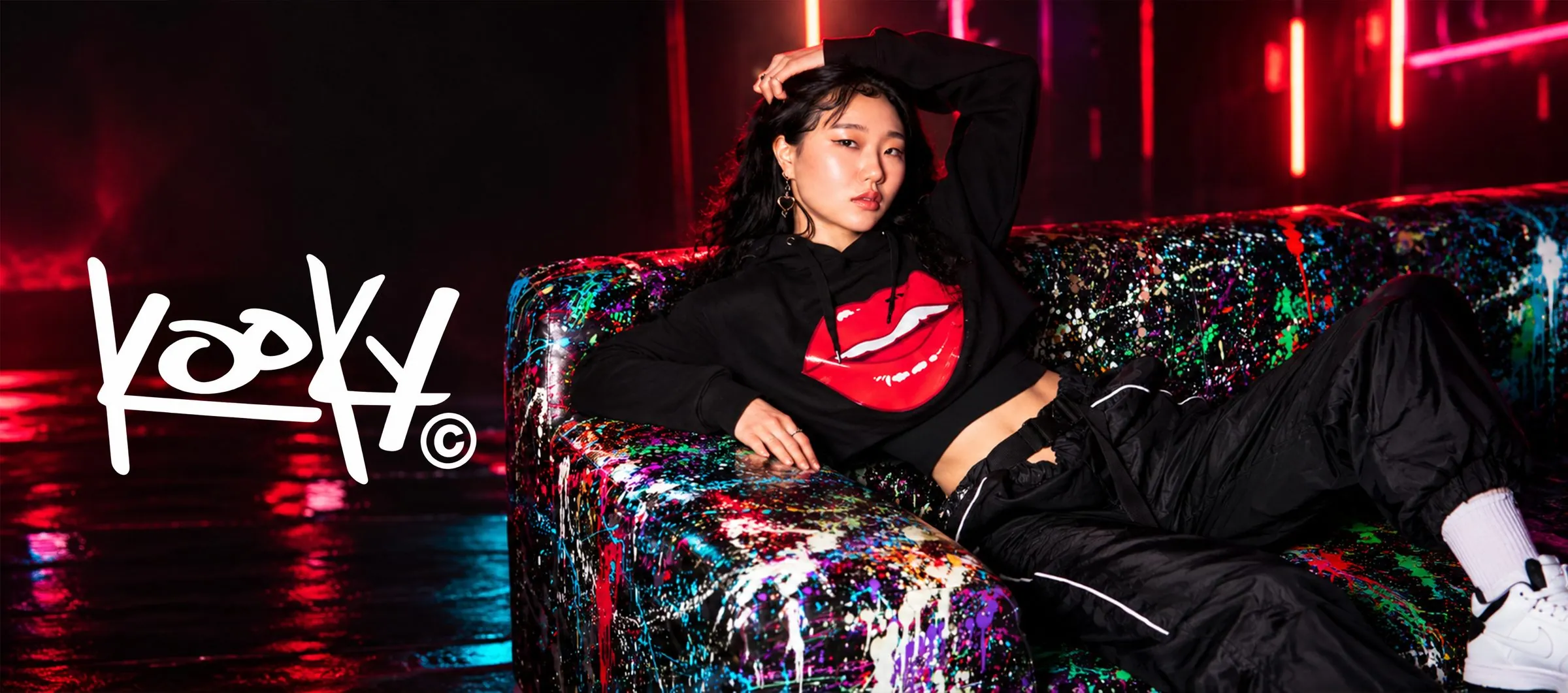 Kooky campaign banner with the Kooky wordmark beside a model in a red-lips hoodie on a paint-splattered sofa under neon lights