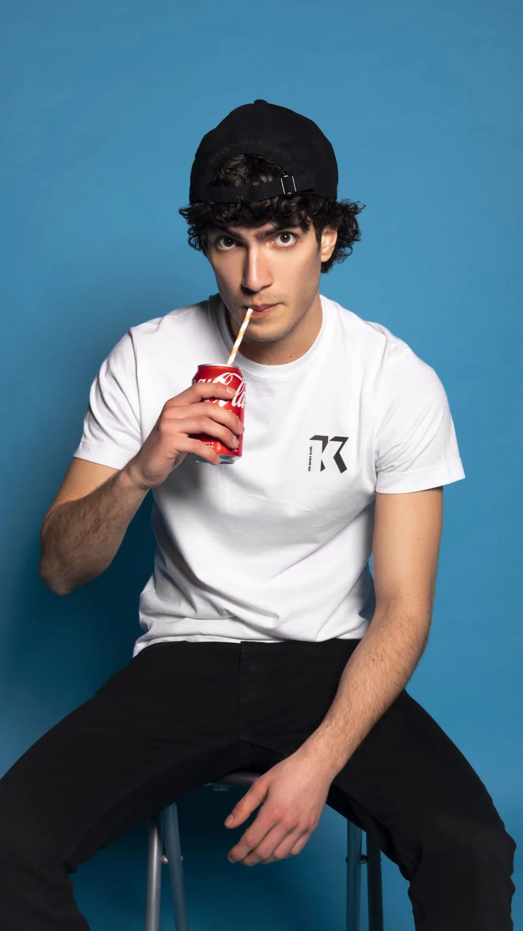 Model in a white Kooky Club T-shirt and cap sipping a drink through a straw against a blue backdrop