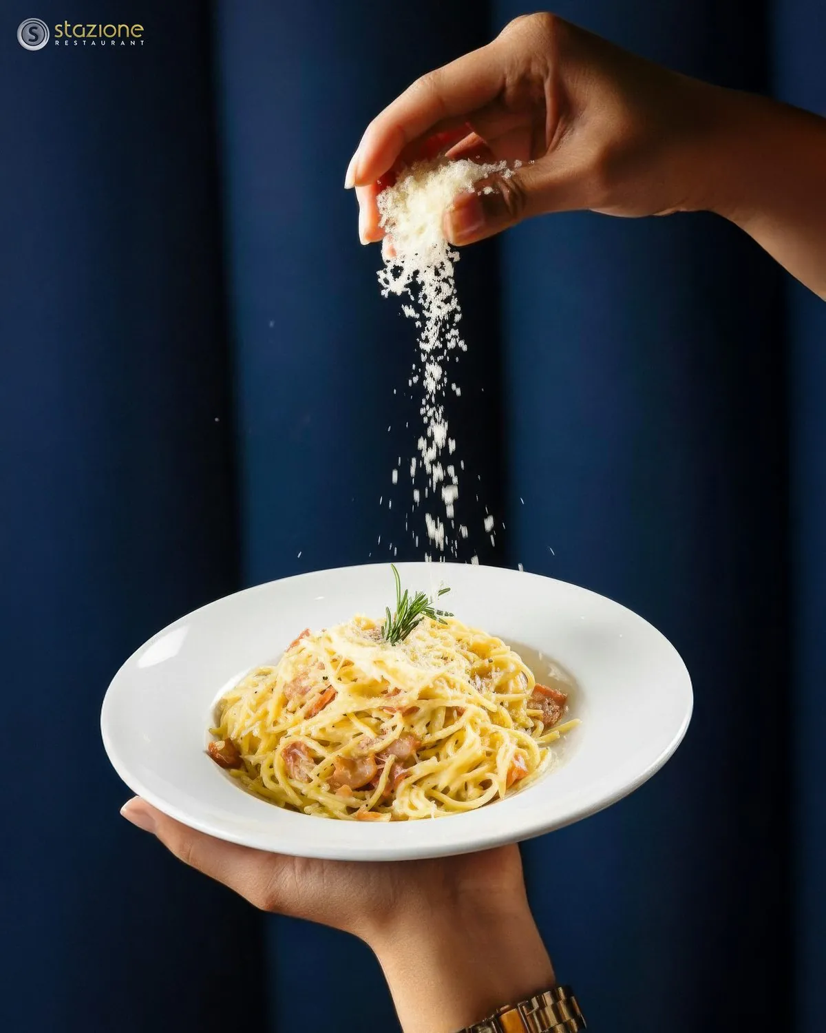 Hand sprinkling parmesan over a plate of spaghetti carbonara against a navy curtain