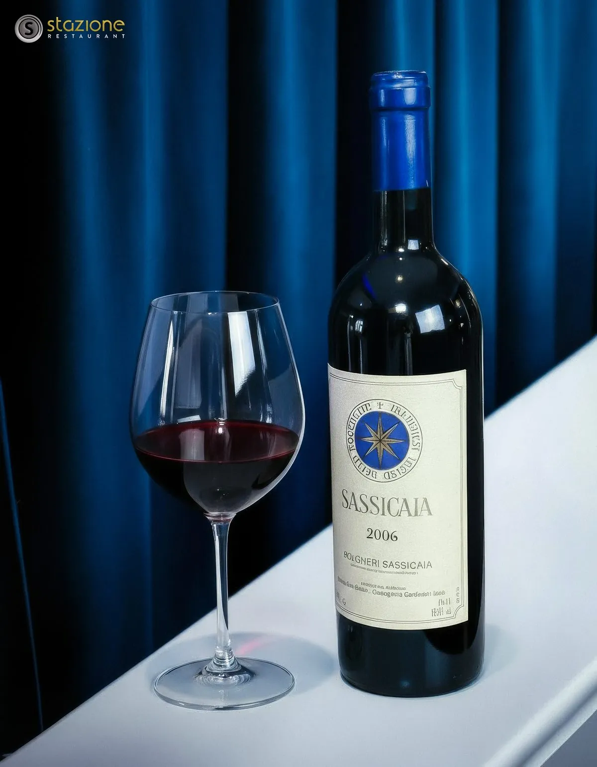 Sassicaia 2006 bottle and a glass of red wine against a deep blue curtain